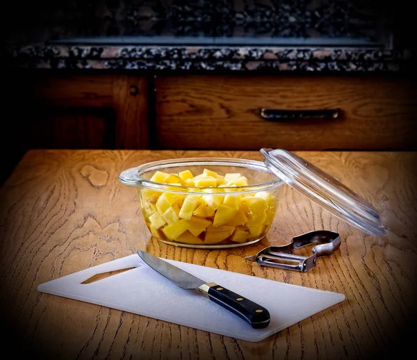 Knife, cutting board, peeler and bowl with cut potatoes on top of the kitchen table