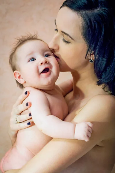 Jovem bela mãe morena sem roupas segurando seus braços e beijando seu filho — Fotografia de Stock