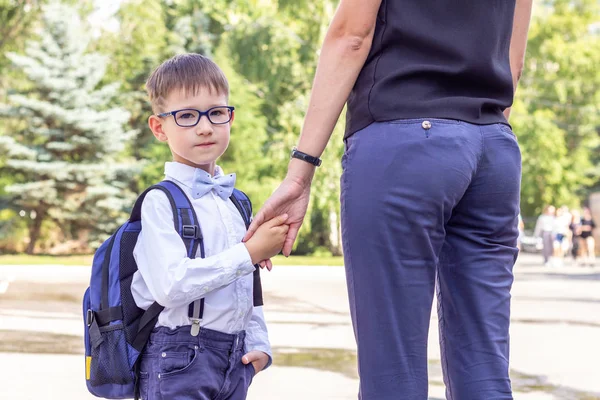 El niño es un niño de primer grado en gafas con un maletín azul sosteniendo su mano madre — Foto de Stock