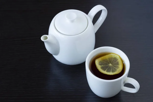 Tea with lemon in a white cup and a white teapot on a dark background — Stock Photo, Image