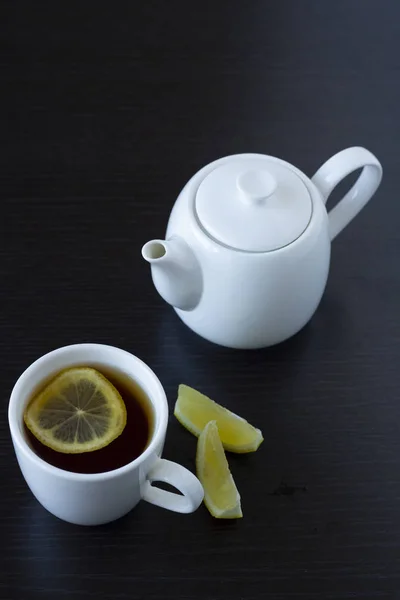 Tea with lemon in a white cup and a white teapot on a dark background — Stock Photo, Image