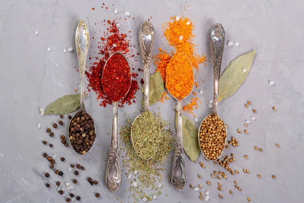 Turmeric powder, paprika, coriander and black peppercorns in metal teaspoons on a gray background