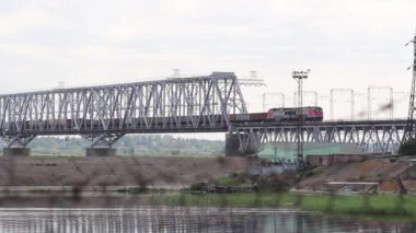 Yazın bulutlu bir havada, yük treni nehir kıyısındaki tren köprüsünden geçer. Yüksek kaliteli FullHD görüntüler