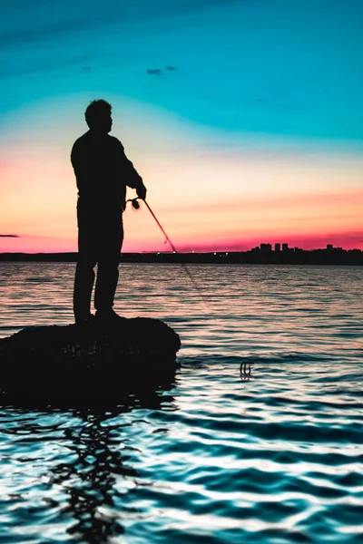 Silhueta de um pescador que fica em uma rocha depois do pôr-do-sol com silhueta da cidade em uma margem do lago — Fotografia de Stock