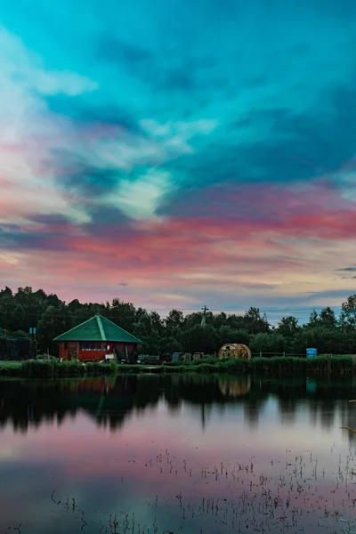 Довгий експозиційний мальовничий вид на маленький червоний будинок на березі ставка, що відображає красиві синьо-рожеві хмари після заходу сонця — стокове фото
