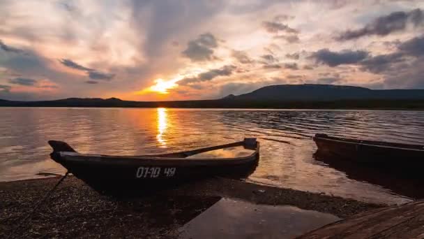 A nap lenyugszik egy hegyi tó felett, és egy csónak horgonyoz a parton. — Stock videók
