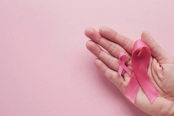 Manos sosteniendo cintas rosas sobre fondo rosa, cáncer de mama awa —  Fotos de Stock