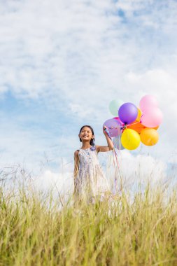 Eller yayılan renkli balonlar çayırda holding mavi gökyüzü ve bulutlar, karşı sevimli küçük kız.