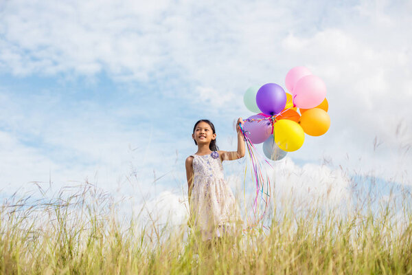 Cute little girl holding colorful balloons in the meadow against blue sky and clouds,spreading hands.