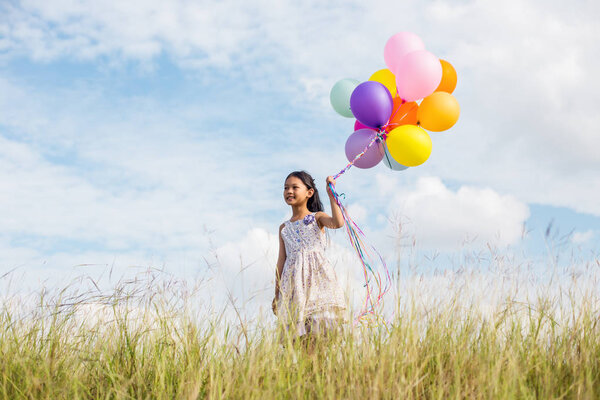 Cute little girl holding colorful balloons in the meadow against blue sky and clouds,spreading hands.