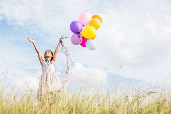 Cute little girl holding colorful balloons in the meadow against blue sky and clouds,spreading hands.