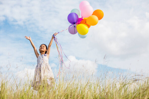 Cute little girl holding colorful balloons in the meadow against blue sky and clouds,spreading hands.