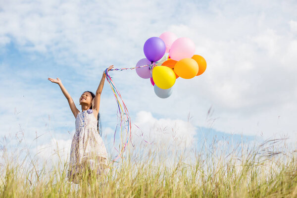 Cute little girl holding colorful balloons in the meadow against blue sky and clouds,spreading hands.