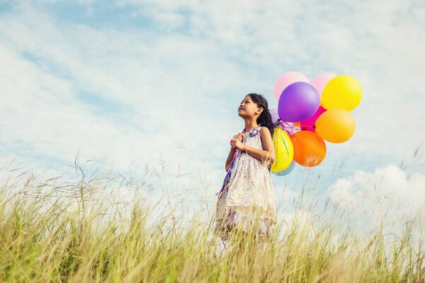 Cute little girl holding colorful balloons in the meadow against blue sky and clouds,spreading hands.