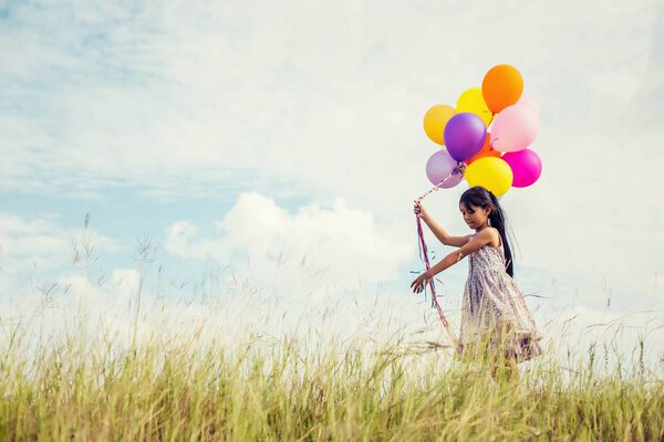 Cute little girl holding colorful balloons in the meadow against blue sky and clouds,spreading hands.