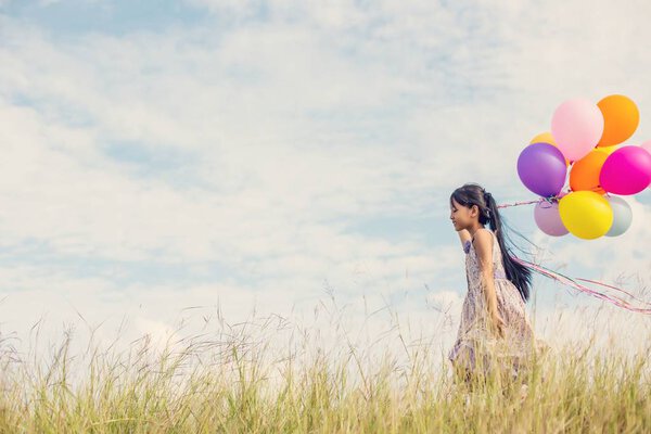 Cute little girl holding colorful balloons in the meadow against blue sky and clouds,spreading hands.