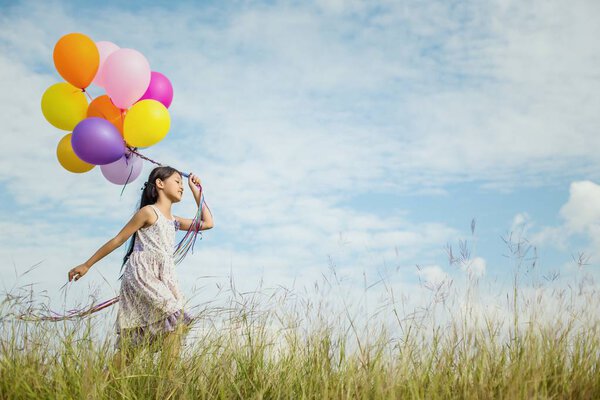 Cute little girl holding colorful balloons in the meadow against blue sky and clouds,spreading hands.