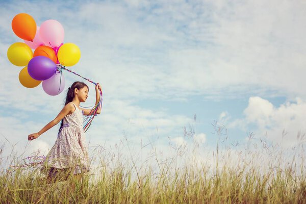 Cute little girl holding colorful balloons in the meadow against blue sky and clouds,spreading hands.