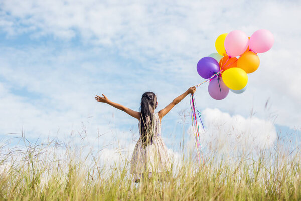 Cute little girl holding colorful balloons, running in the meadow against blue sky and clouds.