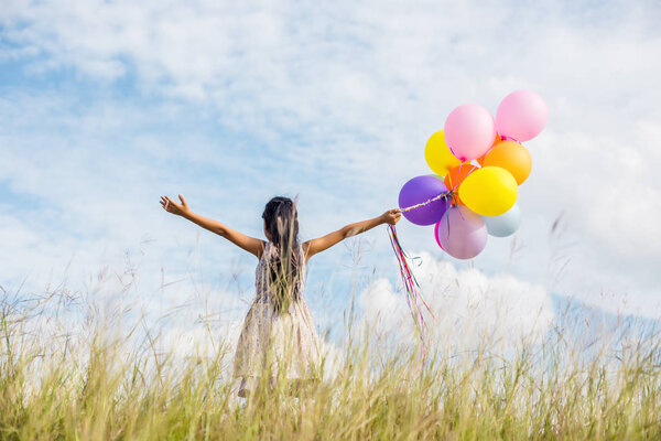Cute little girl holding colorful balloons, running in the meadow against blue sky and clouds.