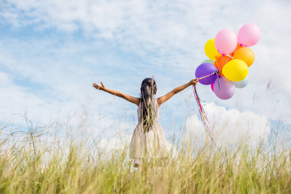 Cute little girl holding colorful balloons, running in the meadow against blue sky and clouds.