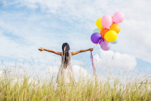 Cute little girl holding colorful balloons, running in the meadow against blue sky and clouds.