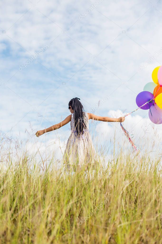 Cute little girl holding colorful balloons, running in the meadow against blue sky and clouds.
