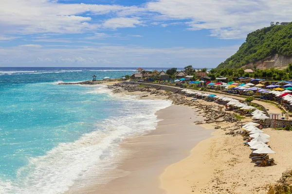 Turistik plaj parlak güneşli bir günde, bir tropik ada sahil şeridi üzerinde. Turkuaz Okyanusu köpüklü dalgalar sarı kum plaj şezlong ve şemsiye ile lots yıkar. Bukit, Bali, Endonezya.