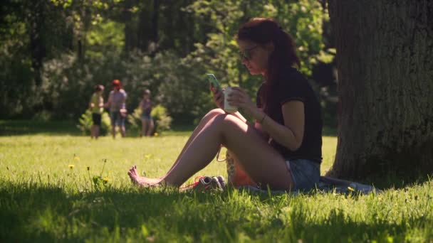 Mujer joven usa teléfono inteligente bebe café en el parque — Vídeo de stock