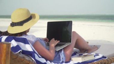 Young woman uses laptop on white beach near ocean