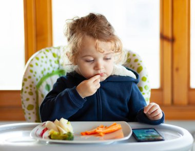 Toddler eats in the high chair while watching movies on the mobile phone. Many children do not want to eat if they do not watch the mobile device or the television. clipart