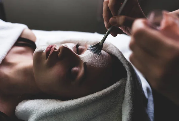 Cosmetician applying facial beauty mask for young beautiful woman at spa salon