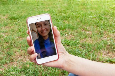 Genç ve gülümseyen bir kızla video görüşmesinde bir akıllı telefonun yakın çekimi. Fotoğraf baharda yeşil bir çimenlikte çekildi..