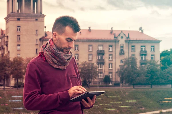 Smart young man works on tablet computer outdoors in urban public space. Millennial using tablet pc with cityscape in background.