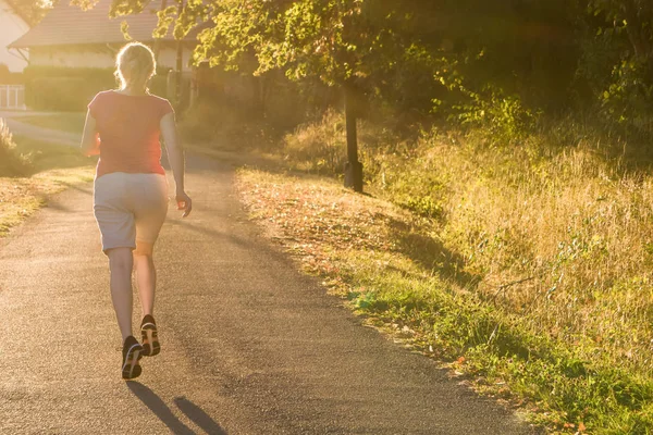Athletic woman running at a countryside trail in sunrise light. Beautiful female running outside in leafy and green suburb.
