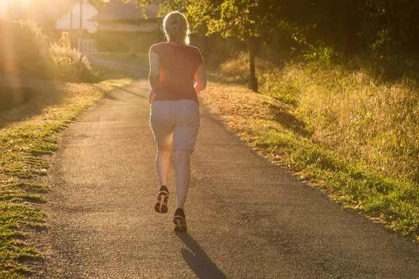 Athletic woman running at a countryside trail in sunrise light. Beautiful female running outside in leafy and green suburb.