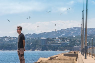 Genç adam limandaki rıhtımda duruyor ve geleceğe bakıyor. Gökyüzünde martılar ve martılar. Akdeniz hattı, Palamos, Costa Brava, İspanya.