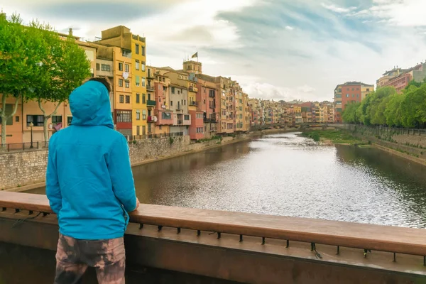 Hombre de chaqueta azul con sudadera con capucha que mira la colorida vista de Girona y el río Onyar desde el puente . — Foto de Stock