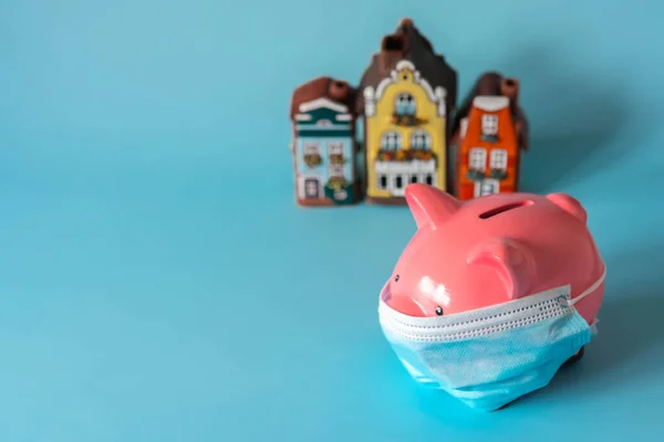 Piggy bank with medical mask and savings for real estate. Piggy bank with house models on blue background. Concept of real estate price during pandemic and quarantine. Selective focus on piggy bank