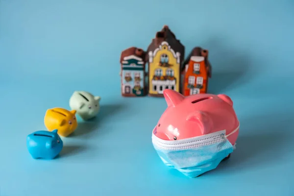 Piggy bank with medical mask, savings for real estate. Piggy bank, small piggy banks and house models on blue background. Concept of real estate price during pandemic and quarantine. Selective focus.