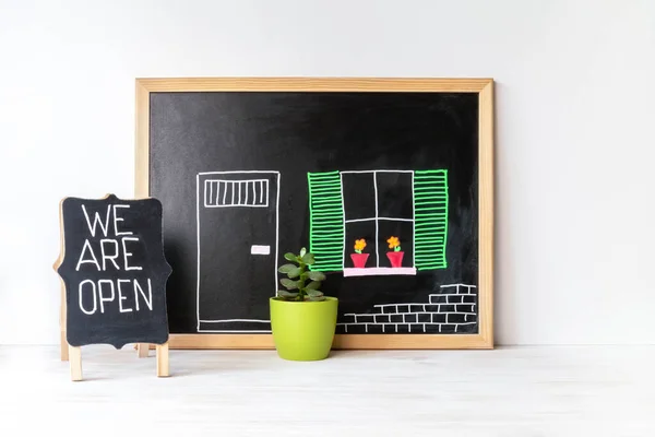 House drawing on blackboard, plant in pot and WE ARE OPEN word on chalkboard. Open singboard near house, drawn on blackboard. Open small business concept