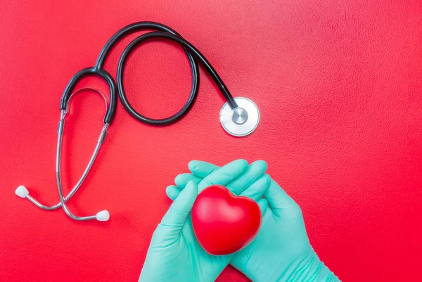 Medical stethoscope and hands in rubber gloves holding red heart on red background. Flat lay of heart in hands and stethoscope. Mockup for medical and health care. Healthcare and cardiology concept. Copy space.