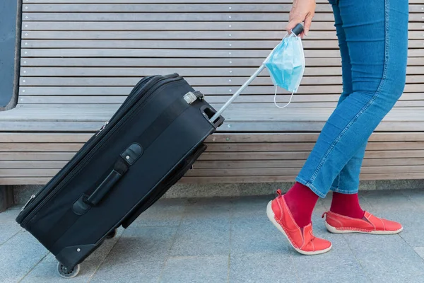 Traveler female holding protective face mask in hand and moving luggage. Woman holding suitcase and disposable medical mask. Legs of woman, mask and baggage. Safe travel during coronavirus