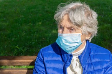 Parkta oturan tıbbi koruyucu maskeli yaşlı kadın. Dışarıda maske takan yaşlı bir kadının portresi. Yaşlılar için virüs koruma kavramı