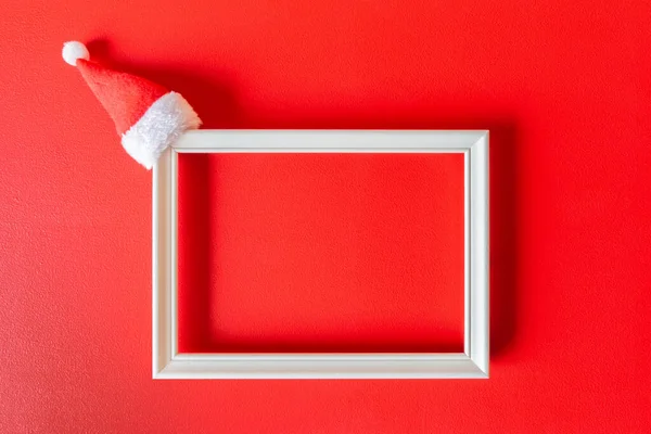 Kırmızı arka planda küçük Noel Baba şapkası olan beyaz çerçeve. Kopya alanı olan minimalist şenlik dekorasyonu. Noel zamanı için maket. Noel, yeni yıl, kış konsepti.