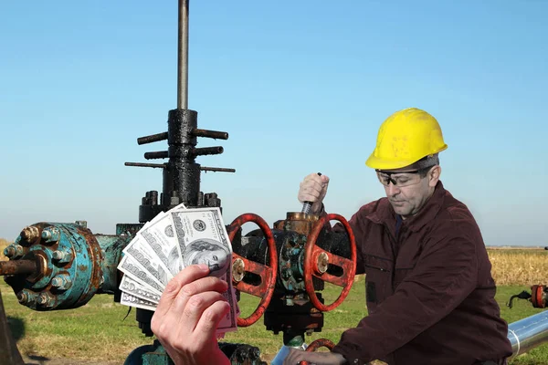石油工人用黄色硬帽子修理阀门在油井与美元票据的手后面 — 图库照片