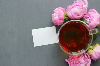 Şeffaf bir fincan sıcak çay, koyu bir arka plan üzerinde pembe şakayık bir buket. Kopya alanı. Metin yazmak için beyaz kart.