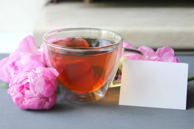 Şeffaf bir fincan sıcak çay, koyu bir arka plan üzerinde pembe şakayık bir buket. Kopya alanı. Metin yazmak için beyaz kart.