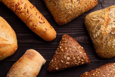 Breads of different types and shapes are spread out on a black background. Rye whole grain bread, wheat unleavened baguettes with sunflower seeds lie on amazing textural background from a natural wood clipart