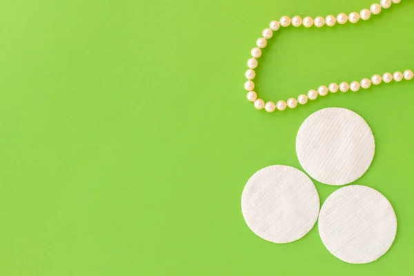 Three wadded disks lie on a green background next to pearl beads. White wadded disks for removal of a make-up. Flat lay. Background for the concept of cosmetology and makeup.